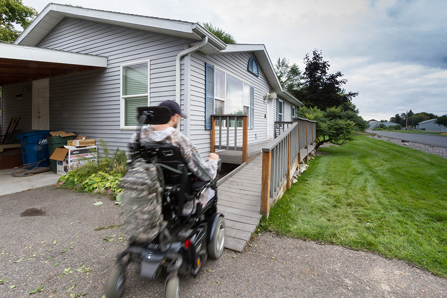 A man wearing a baseball cap uses a power wheelchair to travel up the front grey ramp toward his front door. A camo-colored bag hangs from the back of the chair.